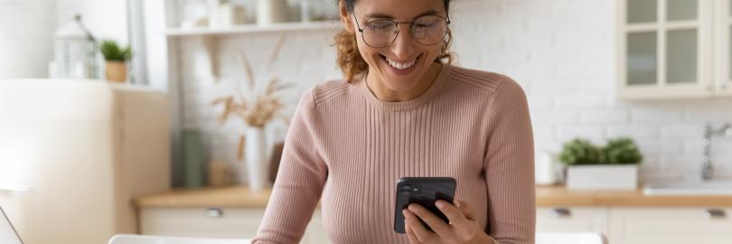 Frau sitzt am Schreibtisch mit vielen Rechnungen, sie schaut auf ihr Handy und tippt in den Taschenrechner