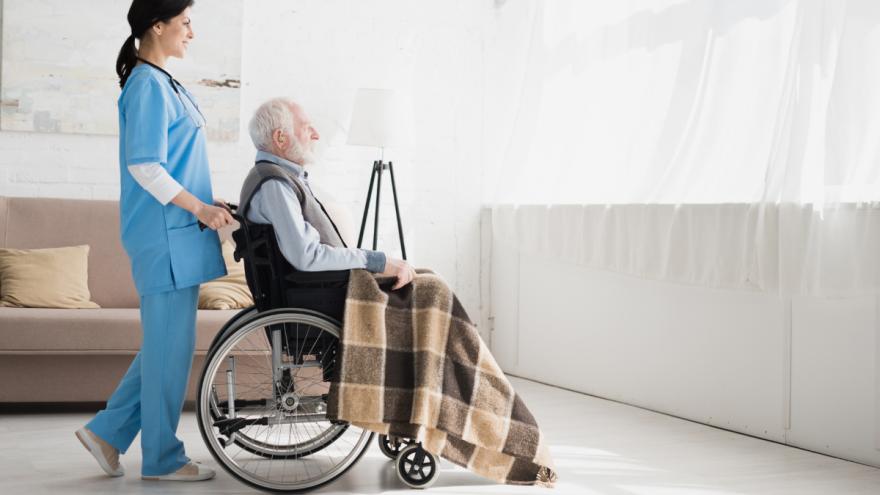 Eine Krankenpflegerin schiebt einen Rollstuhl, in dem ein älterer Mann sitzt.