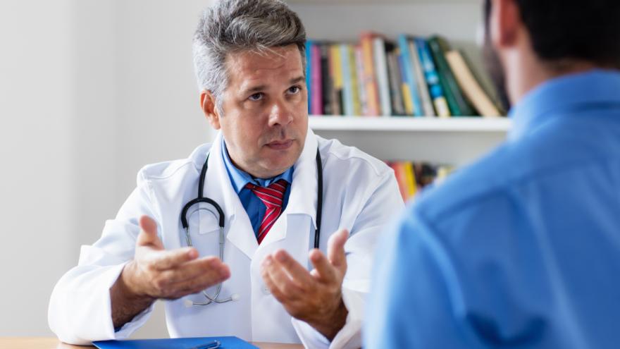 Ein Hausarzt sitzt am Tisch und spricht mit einem Patienten, von dem das Bild nur den Rücken zeigt.