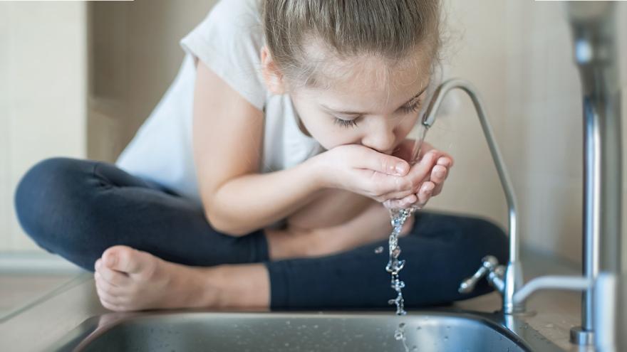 Ein Mädchen sitzt an einem Waschbecken und trinkt Wasser aus dem Hahn.