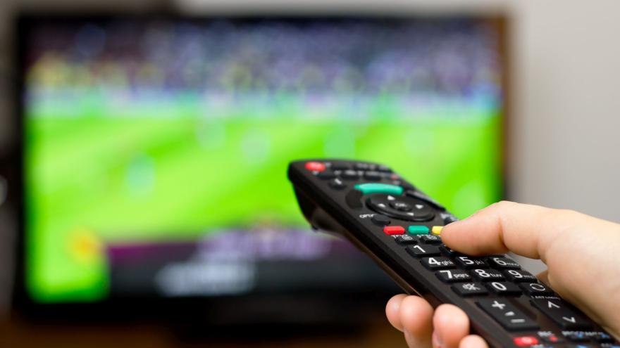 Foto von einer Hand, die eine Fernbedienung hält und auf einen Fernsehbildschirm richtet, auf dem ein Fußballspiel läuft