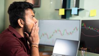 Mensch sitzt vor Computerbildschirmen mit Trading-Kurven und ist schockiert