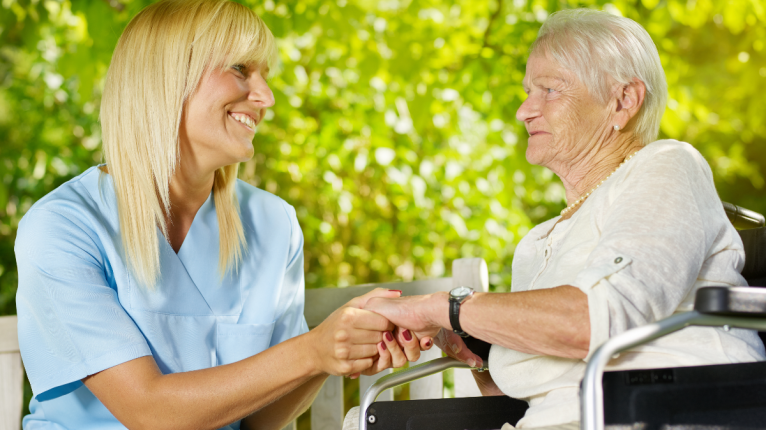 Eine Pflegerin sitzt mit einer älteren Dame draußen und hält ihre Hand.